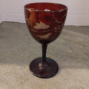 May include: A burgundy-colored wine glass with a stem and a round base. The glass features etched white cloud and bird designs around the bowl. The glass is made of a transparent material.