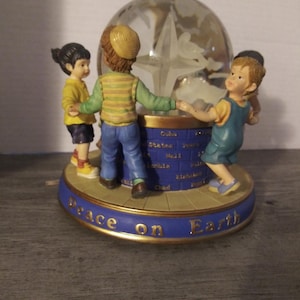 May include: A snow globe with three children holding hands around a globe. The globe is surrounded by a blue base with the words "Peace on Earth" written on it.