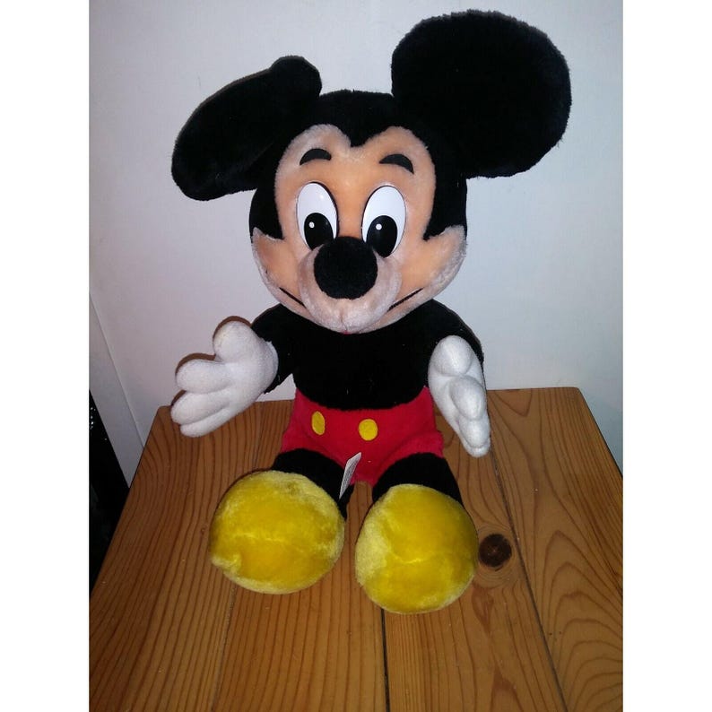 Op de afbeelding: Een pluche Mickey Mouse speelgoed met zwarte vacht, grote oren en een rode en gele outfit. Het speelgoed zit op een houten oppervlak.