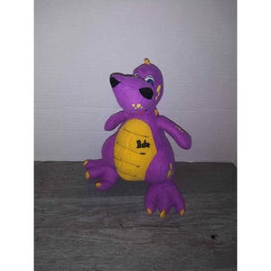 May include: A purple plush dinosaur toy with a yellow belly and accents. The dinosaur has black eyes, yellow spots, and the name "Duke" embroidered on its belly. The toy is standing on a wooden surface.