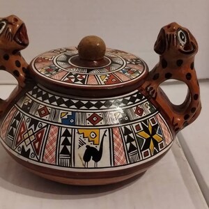 May include: A brown ceramic teapot with a lid and two handles shaped like stylized animal heads. The teapot is decorated with geometric patterns in red, yellow, blue, and white.
