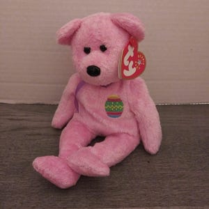 May include: Pink Beanie Baby plush toy with a purple ribbon and an Easter egg design. The tag reads "Beanie Baby" and has a red heart with a white star.