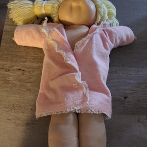 May include: A Cabbage Patch Kid doll wearing a pink robe with white lace trim. The doll has blonde hair and a rosy complexion.