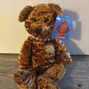 May include: A brown plush teddy bear wearing a pearl necklace. The bear has a gold tag with the word "Happiness" and a red tag with Chinese characters. The tag also says "Fiesta".