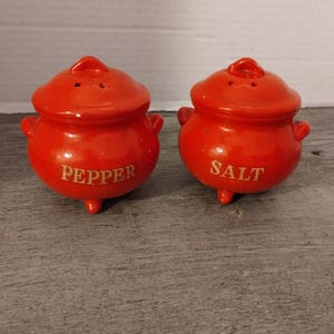 May include: A set of two red ceramic salt and pepper shakers shaped like small pots with lids. The shakers have gold lettering that reads "SALT" and "PEPPER".