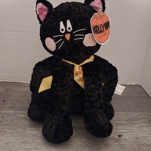 May include: A black plush cat with pink inner ears, white whiskers, and a yellow ribbon. The cat has a tag that says "KELLY HALLOWEEN" with a pumpkin graphic. The cat is sitting on a gray wooden surface.