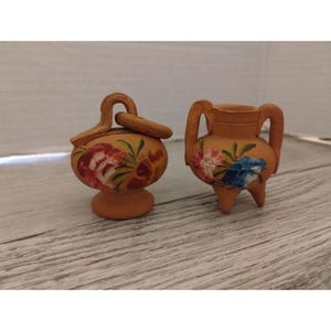 May include: Two small, terracotta-colored ceramic vases. One has a lid and a handle, the other has three legs and two handles. Both are decorated with hand-painted floral designs in red, blue, and yellow.