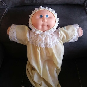 May include: A vintage Cabbage Patch Kids doll with blue eyes and a round face, wearing a yellow and white floral print gown and bonnet with lace trim. The doll's arms are outstretched.