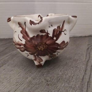 May include: A white ceramic bowl with brown floral designs. The bowl has a scalloped rim and three small feet.