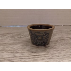 May include: A small, white ceramic pot with a blue floral design. The pot has a brown rim and is empty.