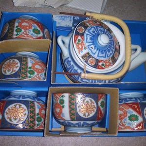 May include: A vintage Japanese tea set with a teapot and four cups, all decorated with floral and geometric patterns in blue, orange, and gold. The teapot has a woven handle. The set is presented in a blue box.