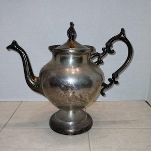 Puede incluir: Una tetera de plata con asa negra y un remate negro en la tapa. La tetera tiene un cuerpo redondo y una base acampanada.