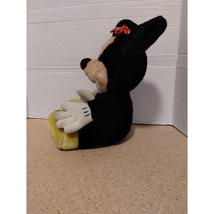 May include: A black and yellow plush toy of Minnie Mouse sitting with her hands in her lap. She has a red bow with white polka dots on her head.