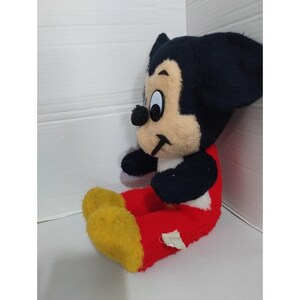 May include: A plush Mickey Mouse toy sitting with red pants and yellow shoes. The toy has black ears and a black nose.
