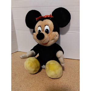 May include: A black and brown plush toy of Mickey Mouse, sitting with a red and white polka dot bow on its head.