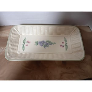 May include: Rectangular ceramic serving tray with a woven basket design. The tray features a cream-colored base with a floral design of purple flowers and pink cherries. The rim is a soft green.