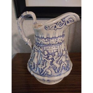 May include: A white ceramic pitcher with blue and white relief designs. The pitcher features a scene of people dancing and playing music. The pitcher has a handle and a spout.