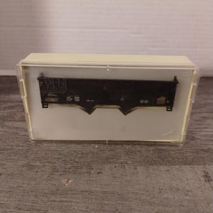 May include: A black model train car in a clear plastic case. The car has the letters "NJ" on the side and the words "CAPY 100000 LIGHT WT 100000" printed on the side.