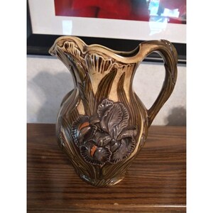 May include: A brown and cream ceramic pitcher with a raised design of an iris flower. The pitcher has a wide mouth and a handle.