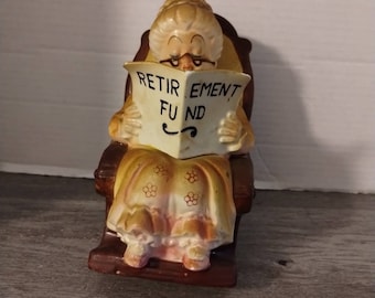Puede incluir: Una figurita de cerámica de una mujer sentada en una mecedora leyendo un periódico que dice "Retirement Fund".