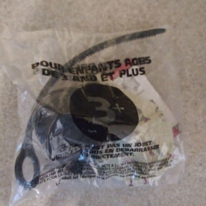 May include: A black toy in a clear plastic bag. The bag has text that reads "POUR ENFANTS AGES DE 3 AND ET PLUS" and a large "3+" symbol. The toy appears to be a spinning top or similar plaything.