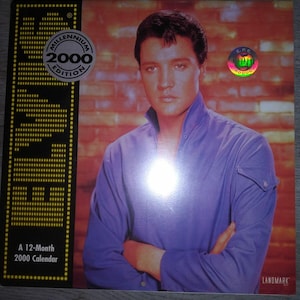 May include: A 12-month 2000 calendar featuring a photo of Elvis Presley in a blue shirt with his arms crossed. The calendar has a black border and a red brick background.