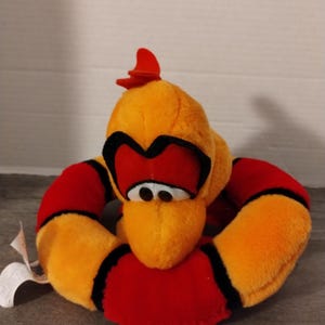 May include: A plush toy snake with a red and orange body and black eyes. The snake is curled into a circle with its head sticking out.