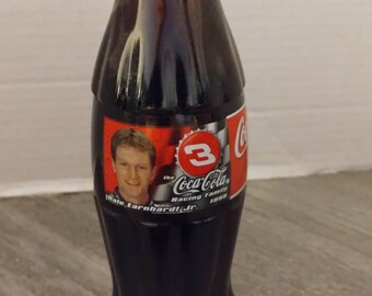 May include: A glass Coca-Cola bottle with a red cap and a label featuring Dale Earnhardt Jr. and the number 3. The label says "The Coca-Cola Racing Family 1999".