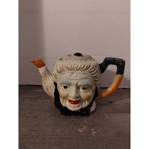 May include: A ceramic teapot shaped like a woman's face with a white, brown, and green glaze. The teapot has a black bow tie and a black and white patterned hat.
