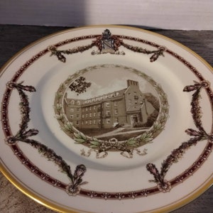 May include: A white ceramic plate with a gold rim and a brown and white image of a building with a wreath around it. The plate has a burgundy and gold border with a floral design.