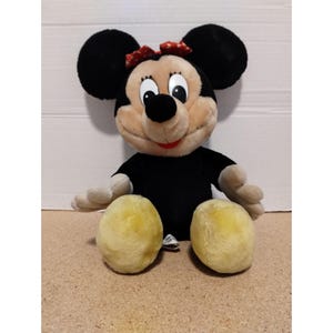 May include: A plush black and yellow Minnie Mouse toy with a red and white polka dot bow. The toy is sitting with its legs crossed and has a friendly expression.