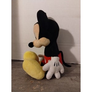 May include: A plush Mickey Mouse toy sitting with his legs crossed. He is wearing his signature red shorts and yellow shoes. His ears are black and his face is brown.