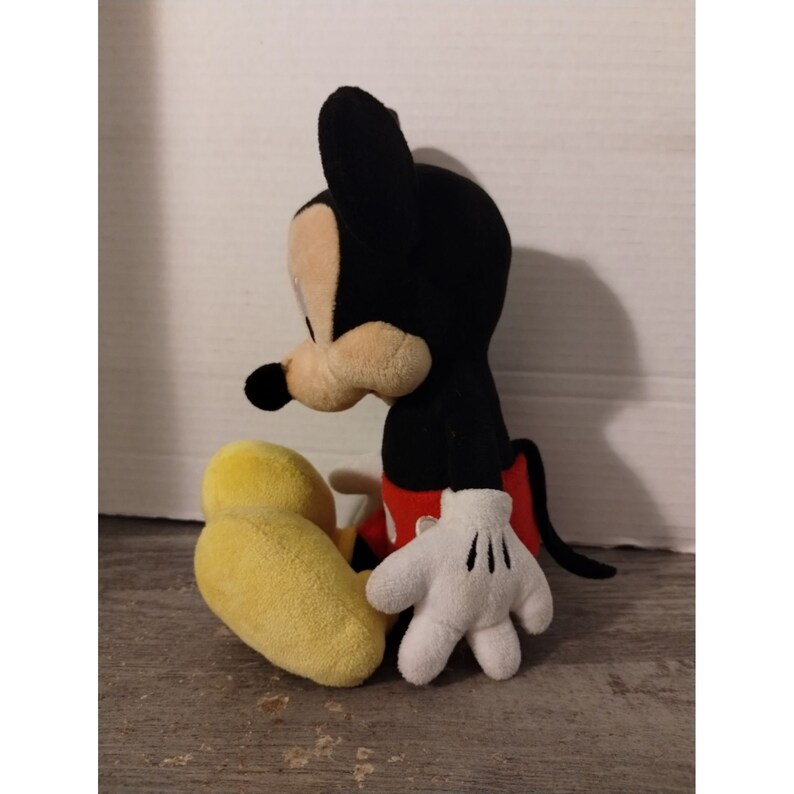 May include: A plush Mickey Mouse toy sitting with his legs crossed. He is wearing his signature red shorts and yellow shoes. His ears are black and his face is brown.