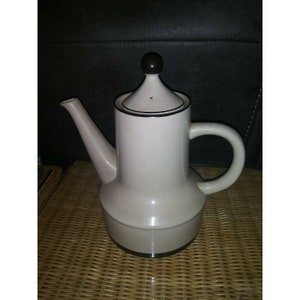 May include: A cream-colored ceramic teapot with a brown lid and a brown knob. The teapot has a wide base and a narrow spout.