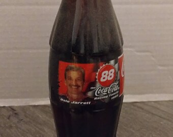May include: A glass bottle of Coca-Cola with a red and white label featuring Dale Jarrett and the text "The Coca-Cola Racing Family 1999". The bottle has a red cap with white text.