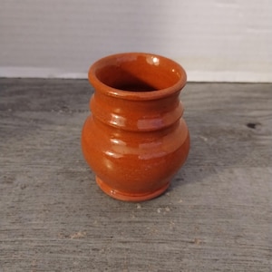May include: A small, handcrafted terracotta-colored ceramic vase. The vase has a rounded body with a narrow neck and a slightly flared rim. The exterior has a smooth, glossy finish. The vase is approximately 4 inches tall.