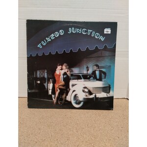 May include: A vintage vinyl record album cover featuring a white convertible car with two women in dresses and a man in a tuxedo standing next to the car. The album cover has the text "TUXEDO JUNCTION" in a neon blue font.