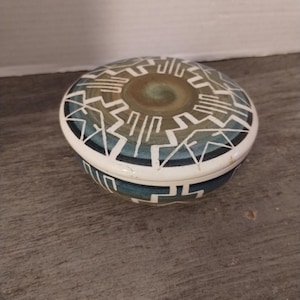 May include: A small, round ceramic trinket box with a lid. The box is decorated with a geometric pattern in shades of blue, brown, and white.