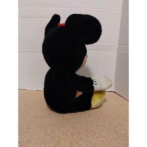 May include: A black plush Mickey Mouse toy sitting with its legs crossed. The toy has a red bow and yellow fabric on its feet.