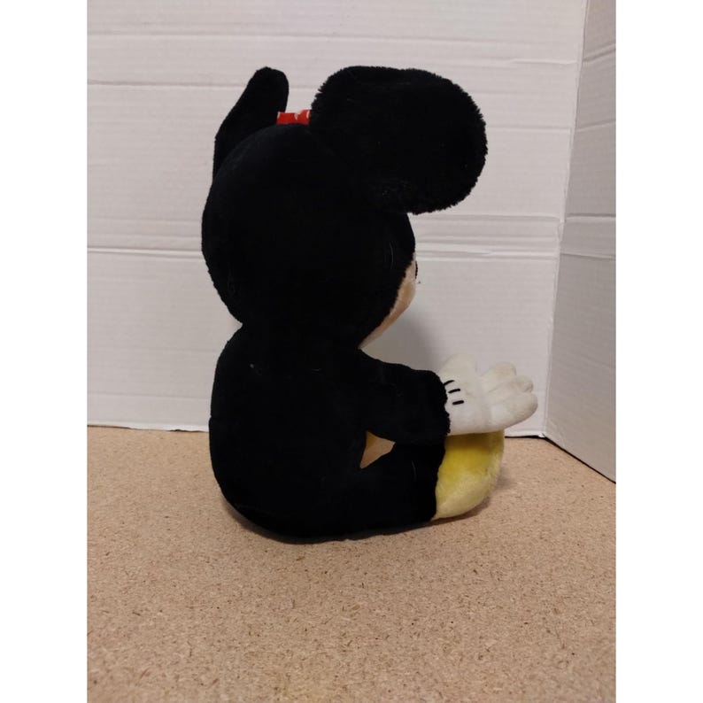May include: A black plush Mickey Mouse toy sitting with its legs crossed. The toy has a red bow and yellow fabric on its feet.
