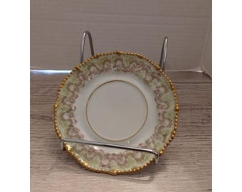 May include: A small, white porcelain plate with a gold rim and a decorative floral pattern in shades of green and pink. The plate is held in a silver metal stand, showcasing its delicate design. The plate is approximately 6 inches in diameter.