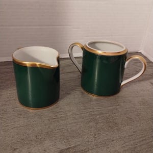 May include: A set of two green and gold creamer and sugar containers. The containers are made of porcelain and have a gold trim around the top.