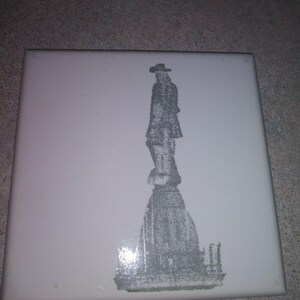 May include: A square, white ceramic tile featuring a gray silhouette of a person in a hat standing atop a monument. The image is a detailed, grayscale illustration, suitable for home decor.