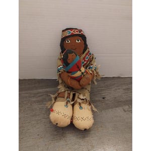 May include: A handmade fabric doll dressed in traditional Native American clothing, holding a smaller doll. The larger doll has brown skin, black hair, and a beaded headband. The smaller doll is wrapped in a colorful blanket.