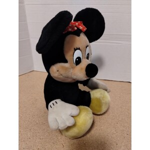 May include: A plush black and tan Minnie Mouse toy with a red polka dot bow. The toy is sitting with its legs crossed and has a big smile.