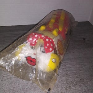 May include: A white plush toy in the shape of a cartoon character with red lips, yellow hair, and a red and white polka dot dress. The toy is wrapped in clear plastic and has a yellow and red striped design on the back.