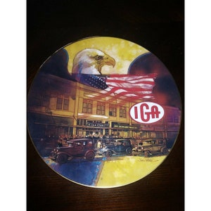 May include: A vintage-style ceramic plate featuring a bald eagle with an American flag, a building with the text "IGA Stores", and a crowd of people in front of the building. The plate has a gold rim and a yellow background.