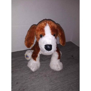 May include: A plush toy dog with brown and white fur, black eyes, and a black nose. The dog has floppy ears and is in a sitting position. The toy has a tag on its left leg.