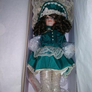 May include: A porcelain doll in a box, wearing a teal dress with white lace trim and a matching bonnet. The doll has brown curly hair, blue eyes, and is packaged in bubble wrap. The doll's shoes are cream-colored.