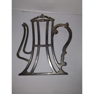 May include: Silver-toned metal teapot-shaped hot pad. The hot pad is a cutout design with a decorative handle.